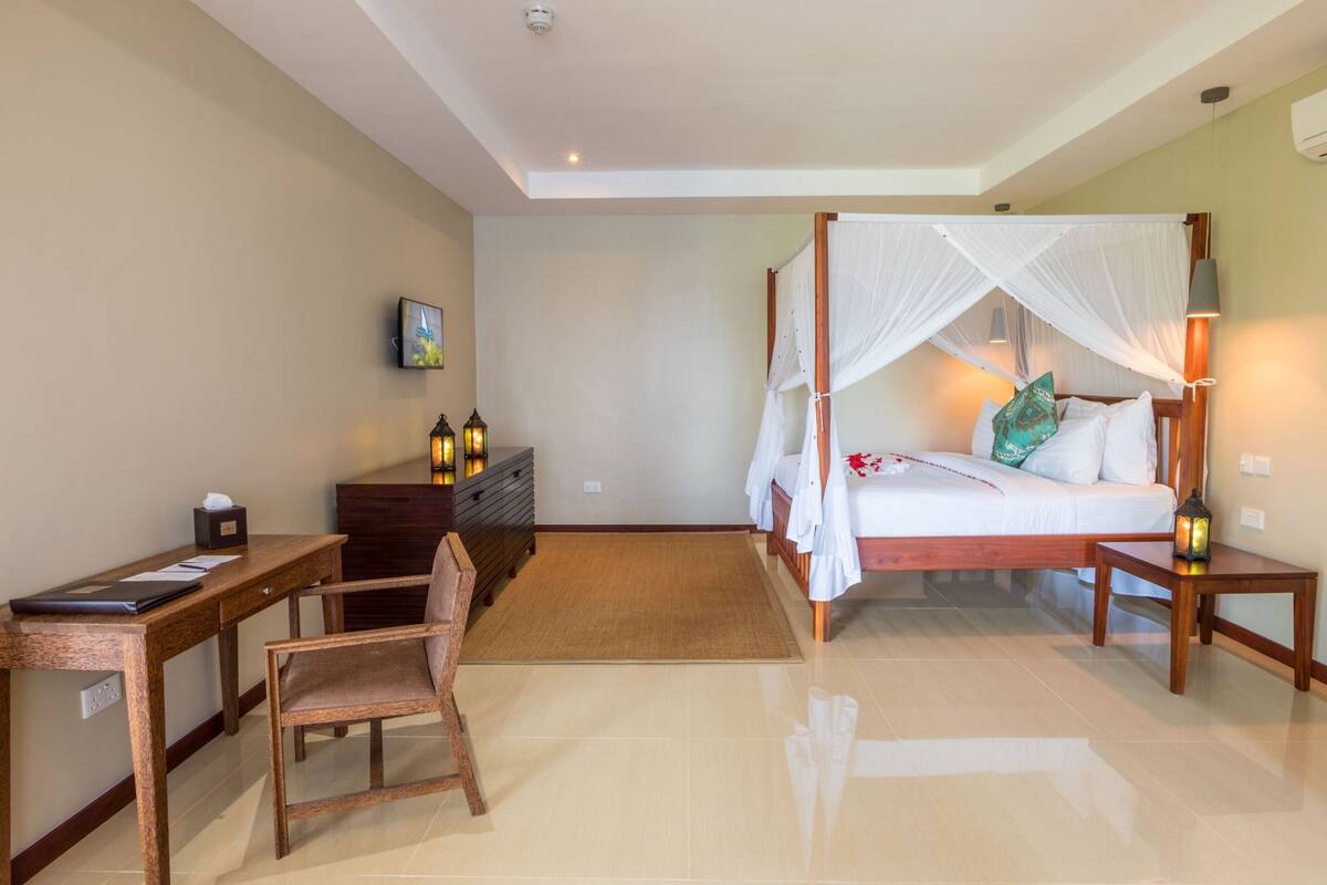 Quarto com cama de dossel, uma escrivaninha, uma cadeira e mesa de cabeceira com abajur.