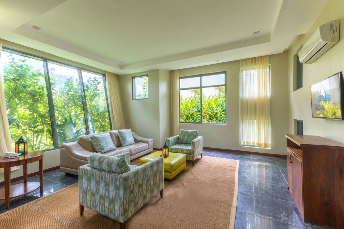 Uma sala de estar ampla com grandes janelas, um sofá, poltronas, uma mesa de centro e um aparador de madeira.