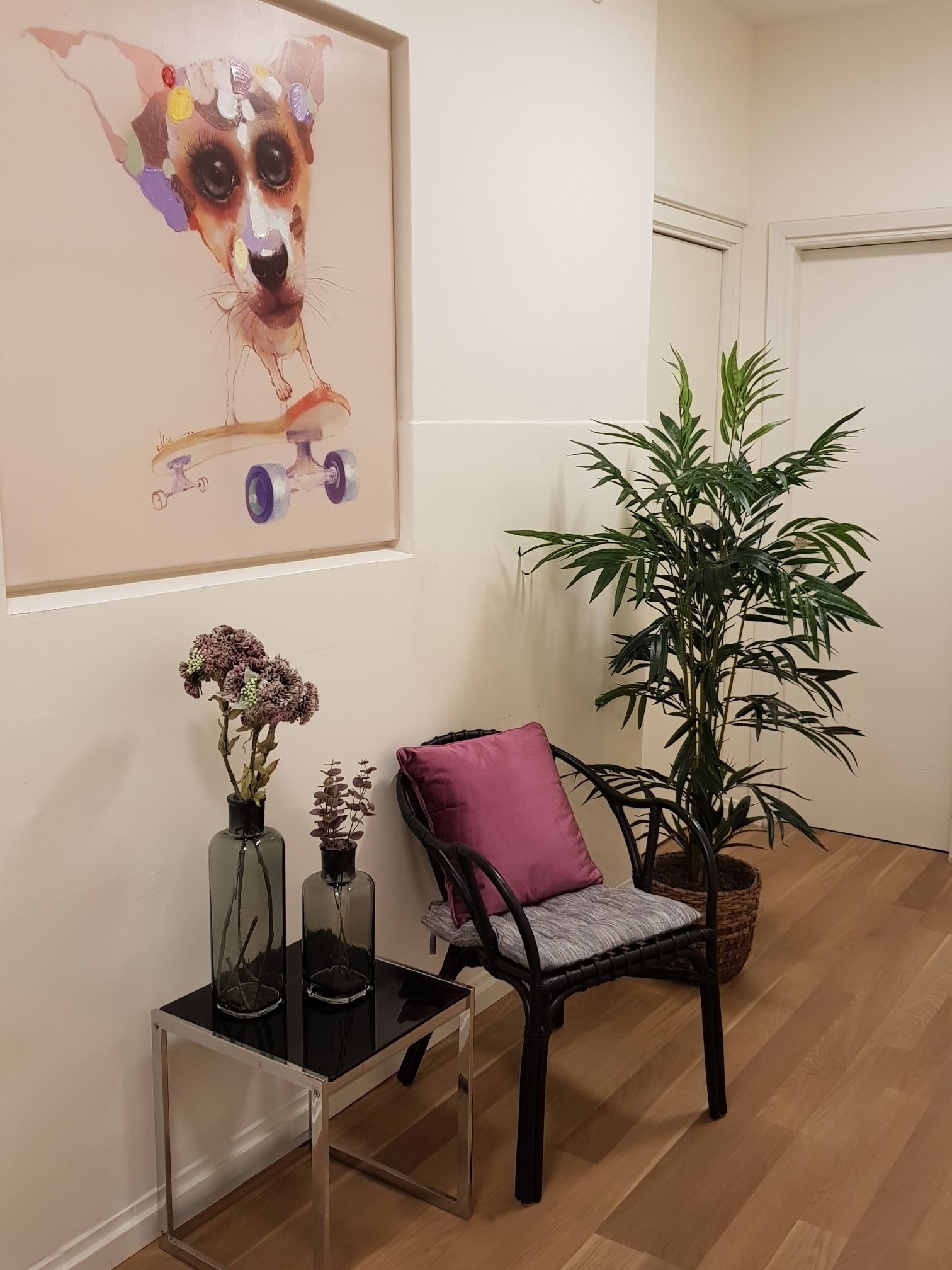 Una habitación con una obra de arte enmarcada de un perro, una silla con un cojín morado, una planta en maceta y una mesita con jarrones.