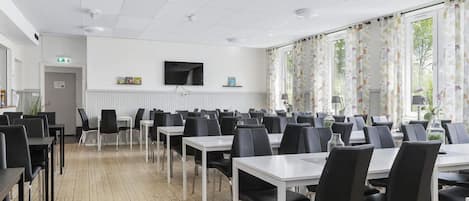 Un salón de conferencias con mesas y sillas, un televisor en la pared y ventanas grandes con cortinas.