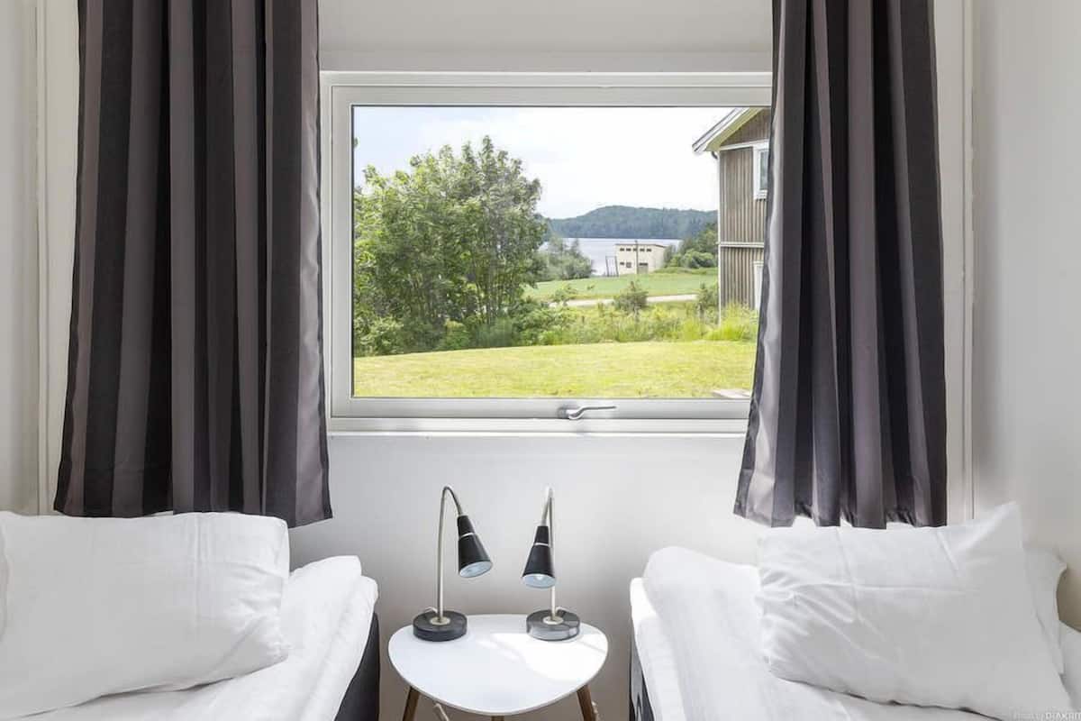 A room with two white beds, a small table with two lamps, and a window with curtains.