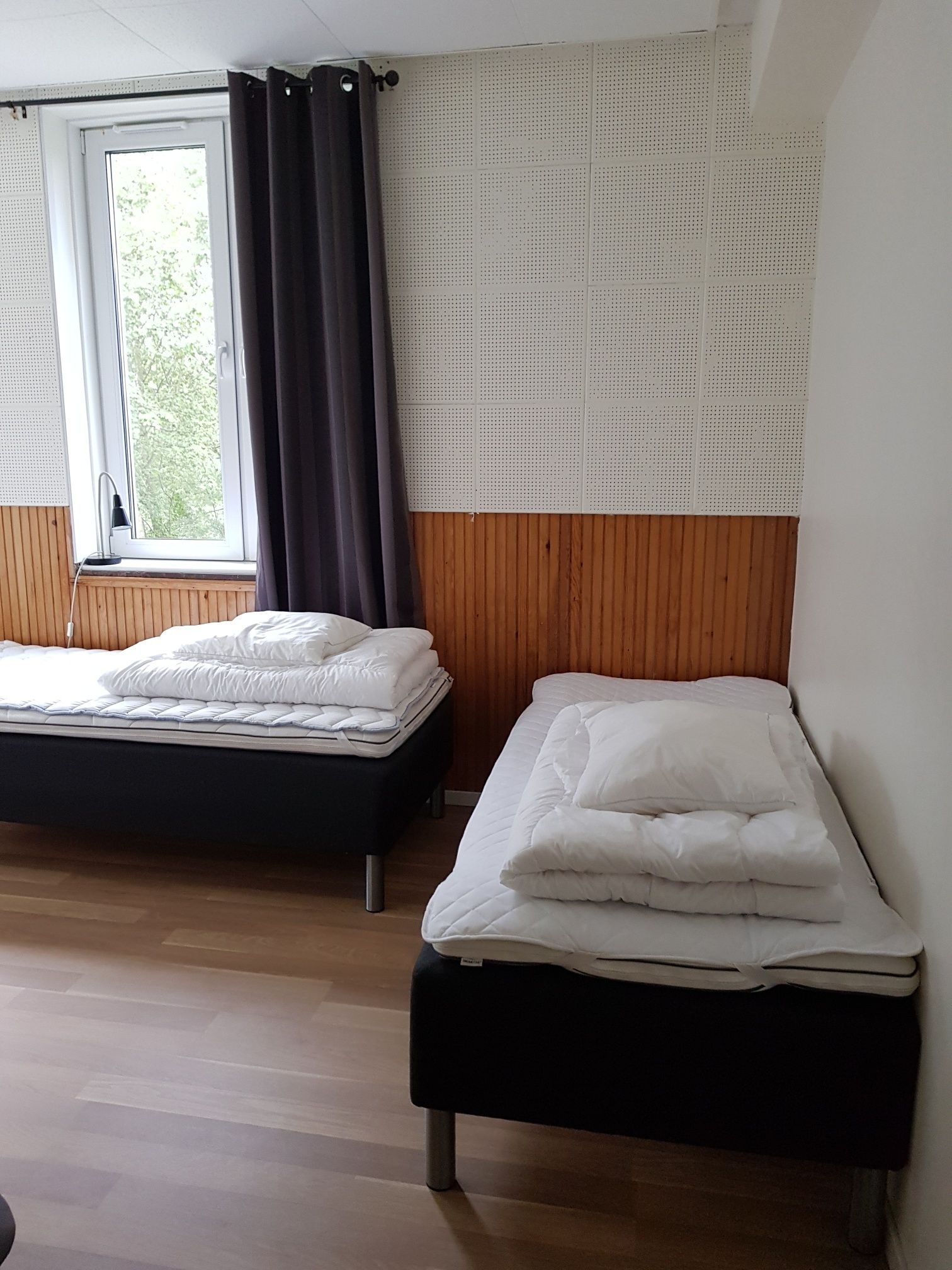 Una habitación con dos camas, una ventana con cortinas, un panel de pared de madera y un techo blanco.