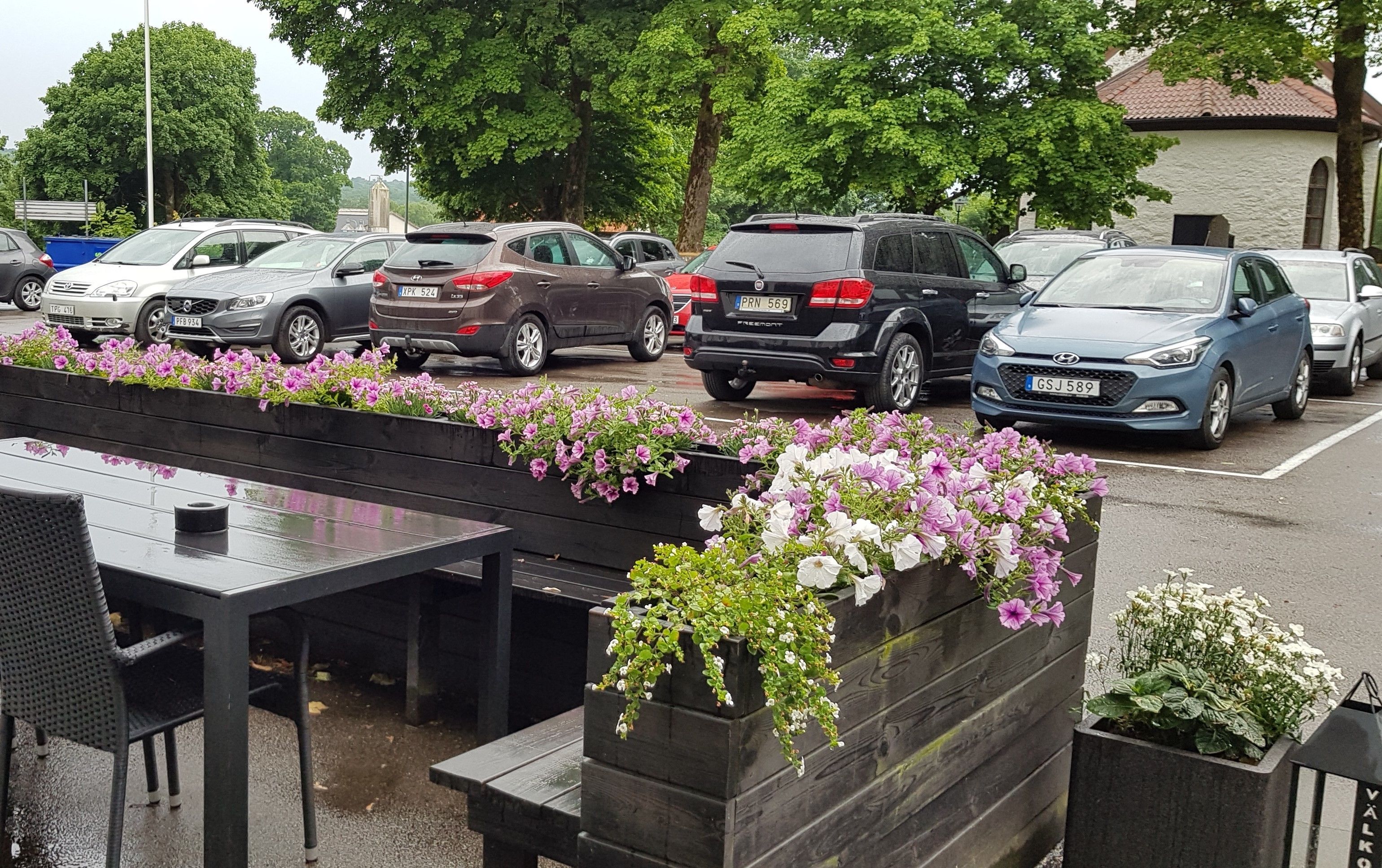 A parking lot with several cars, a table with chairs, and flower planters with pink and white flowers.