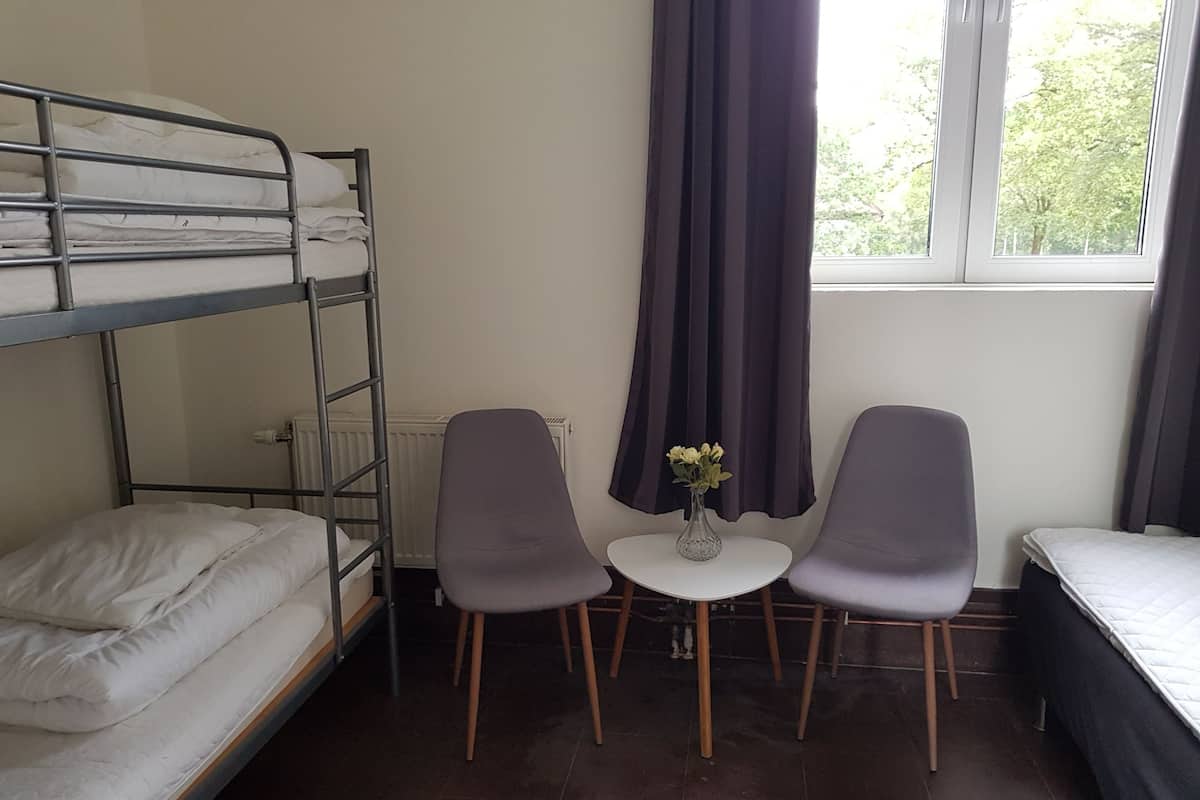 A dormitory room with bunk beds, a small table with chairs, and a window with curtains.