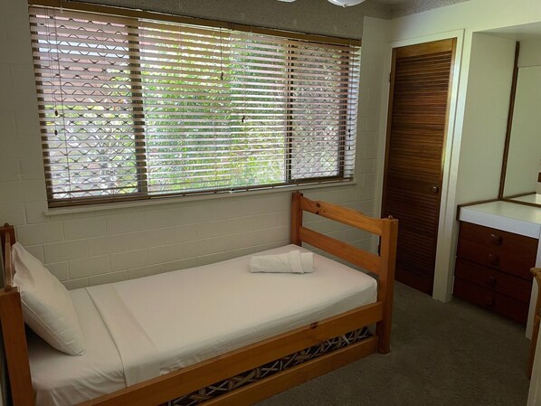 A single bed with a wooden frame and white bedding, a window with blinds, a wooden door, and a small dresser with drawers. - Riverview Two (Mooloolaba)