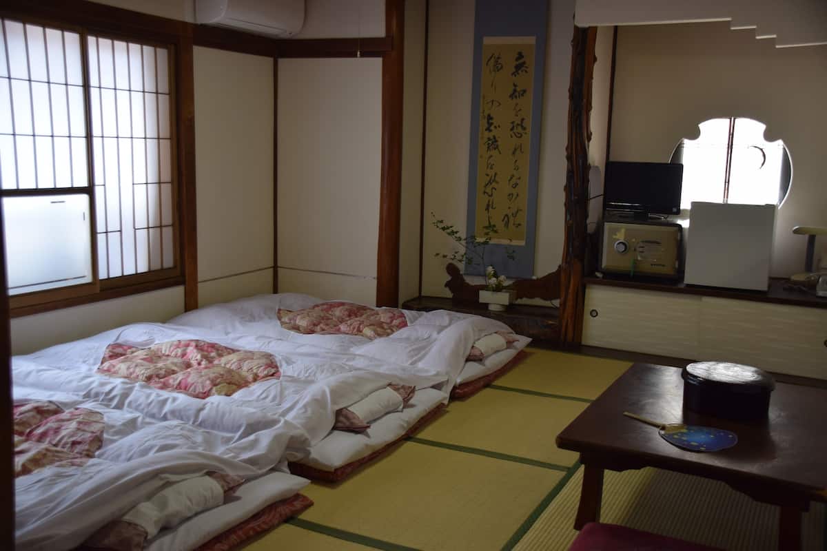 A traditional Japanese room with tatami flooring, a futon bed, a small table with a bowl and chopsticks, and a television on a stand.