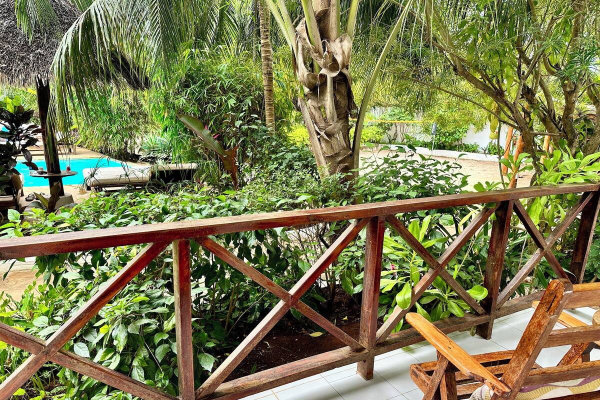 Eine Holzterrasse mit Metallgeländer, umgeben von üppiger Vegetation und einem Pool im Hintergrund.