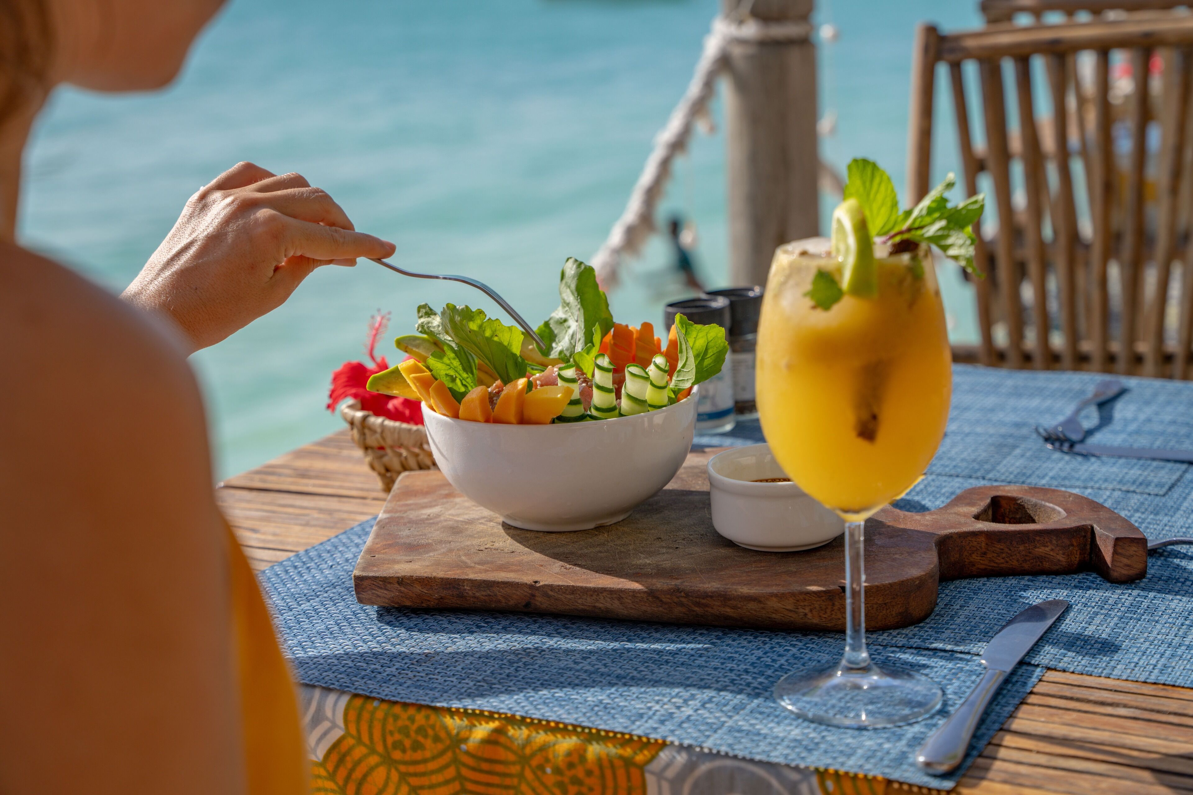 A dining table with a salad bowl and a glass of yellow drink, set on a wooden board with a blue tablecloth.