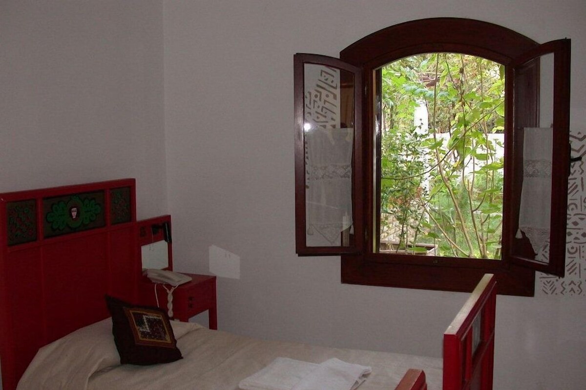 Una habitación con cama, una mesita de noche, un ventanal con contraventanas abiertas y vistas al exterior.