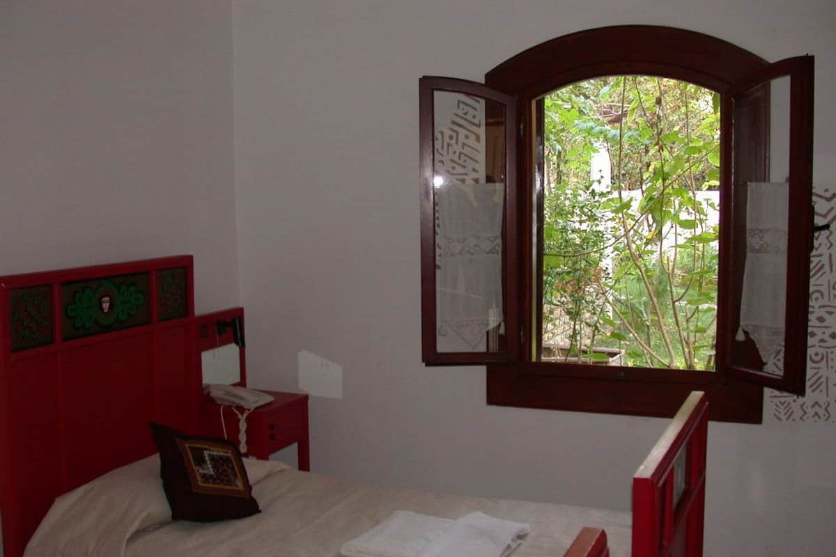 Una habitación con cama, mesita de noche, cabecera roja, ventana con persiana abierta y vista a espacios verdes.