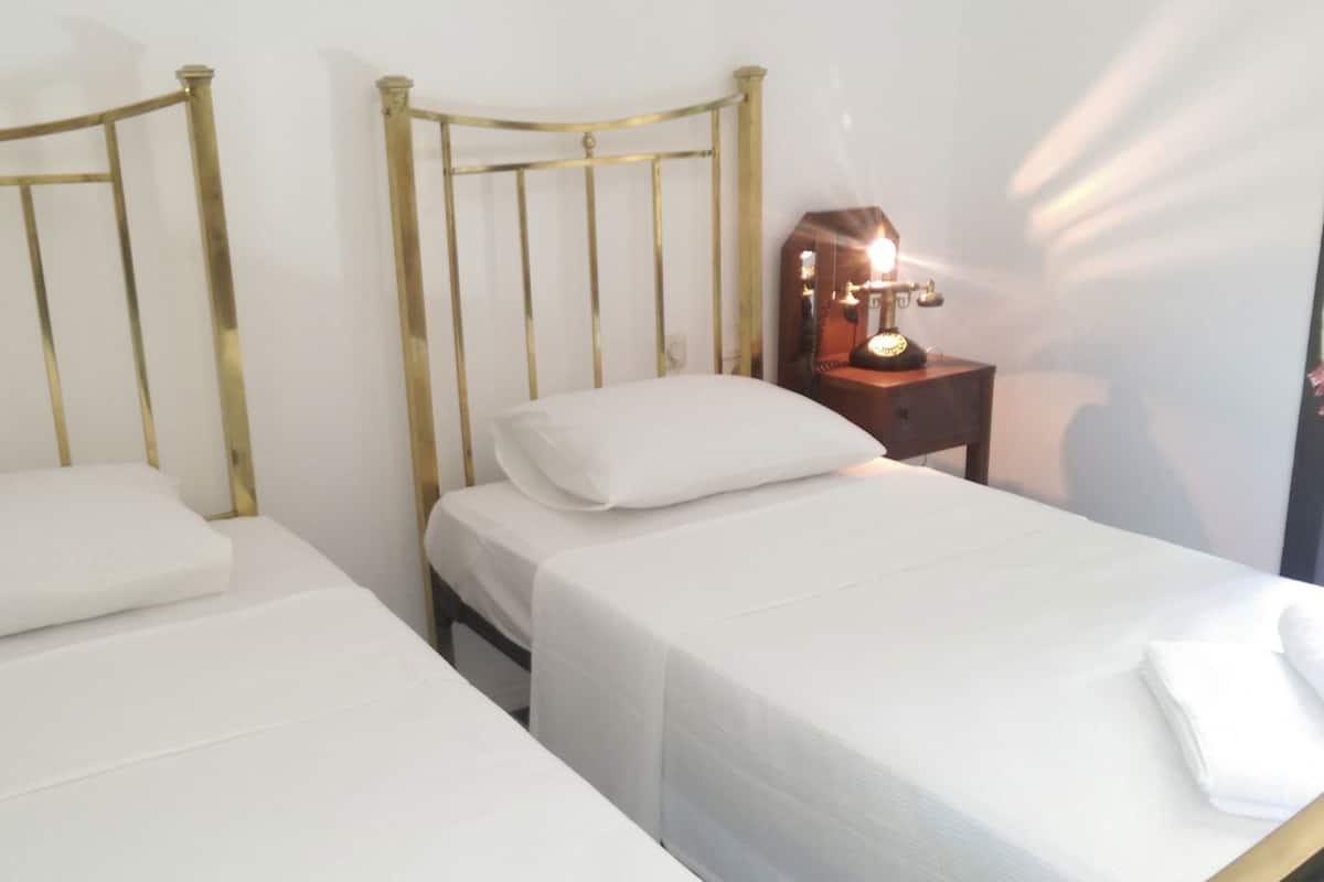 Dos camas individuales con estructura metálica y ropa de cama blanca, una mesita de noche con lámpara y un enchufe en la pared.