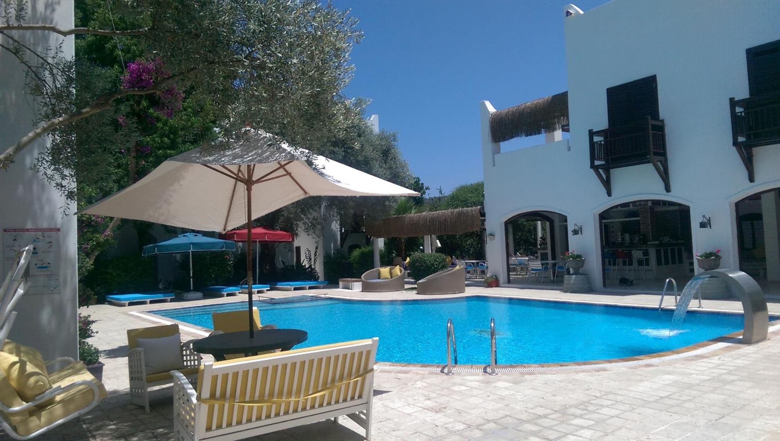 A swimming pool with lounge chairs and umbrellas, a building with arched windows, and a water feature.