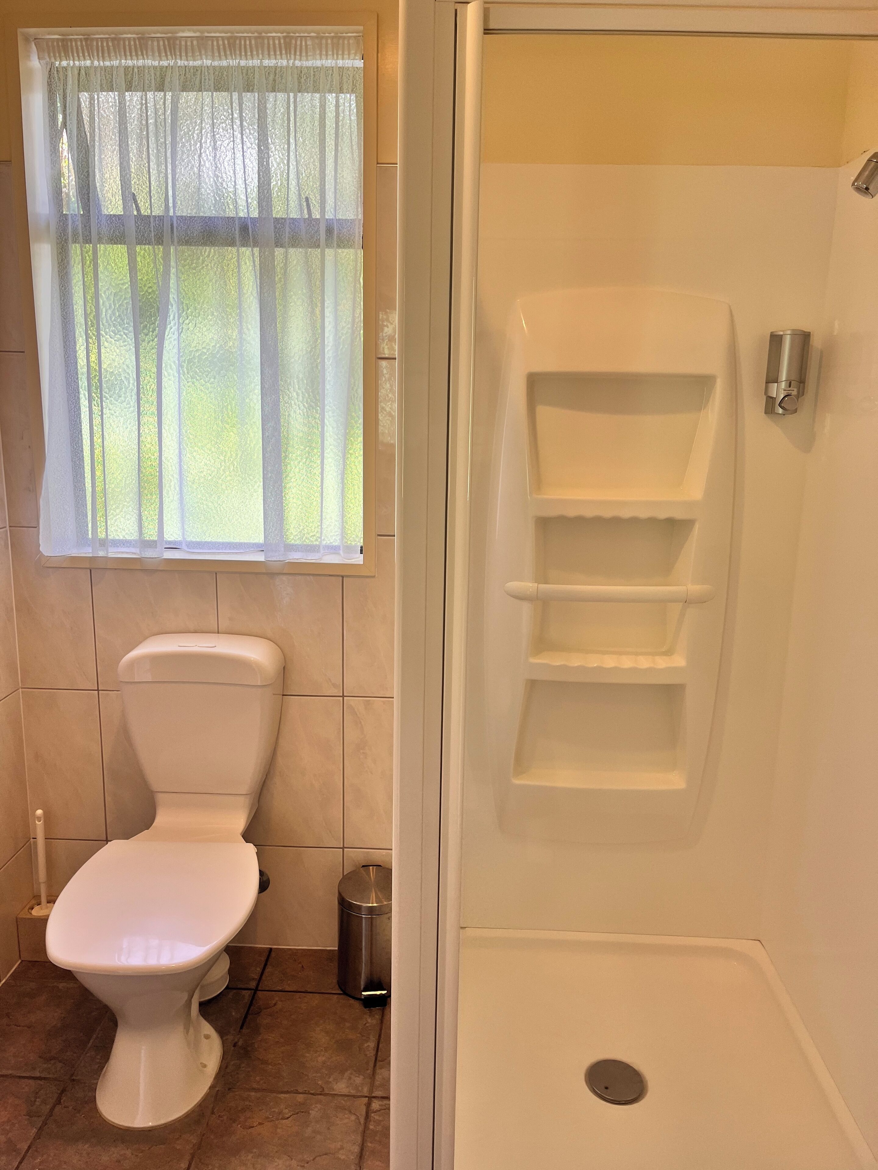 Un baño con inodoro, una ventana con cortina translúcida y una cabina de ducha con estantes integrados.