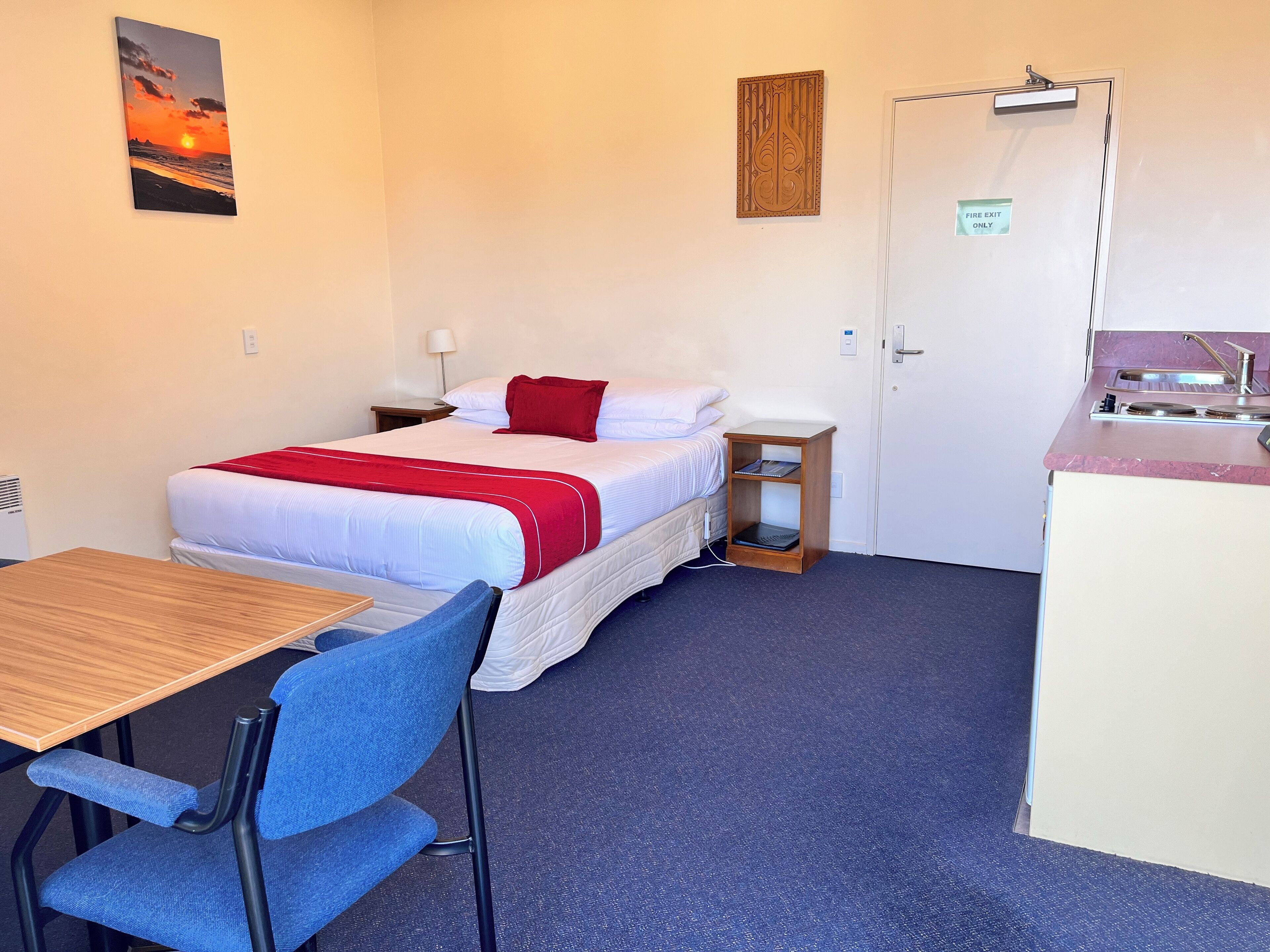 Habitación de hotel con cama, escritorio, silla, pequeña mesa, mesita de noche, puerta y una obra de arte colgada en la pared.