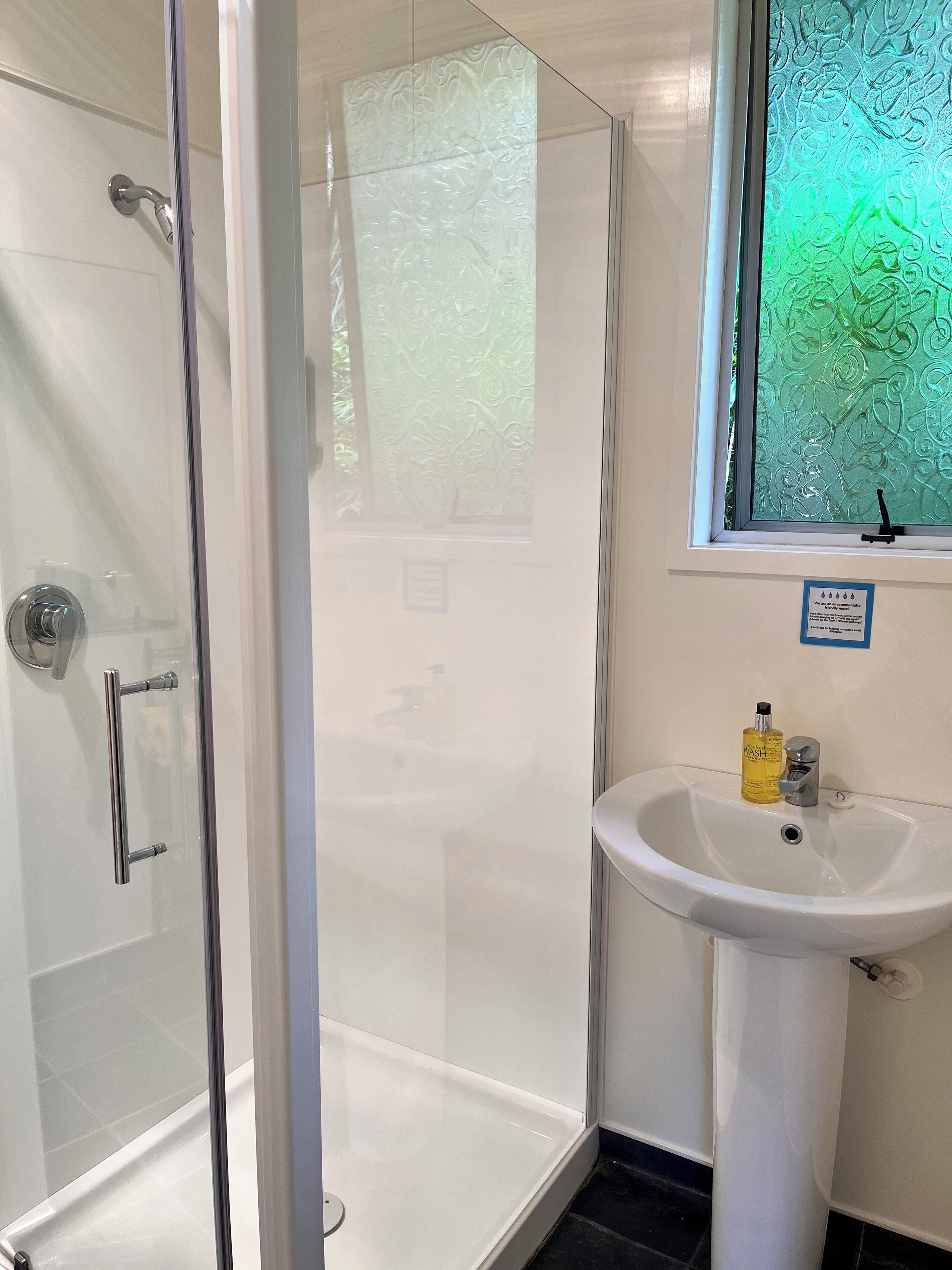 Une salle de bain avec une douche à parois de verre, un lavabo blanc avec un savon, et une fenêtre ornée d’un motif décoratif.