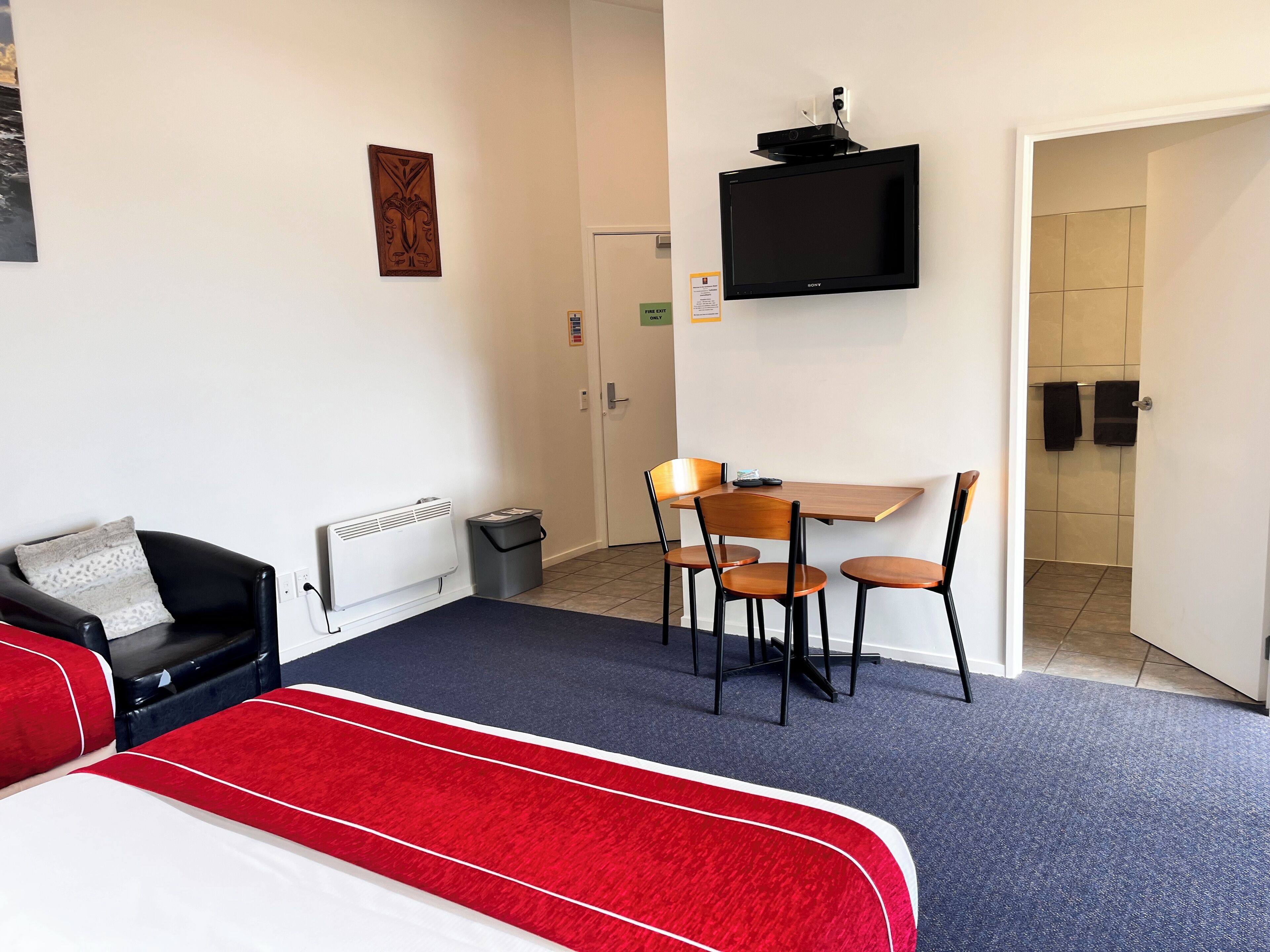 A hotel room with a bed, a desk with chairs, a TV, and a radiator.