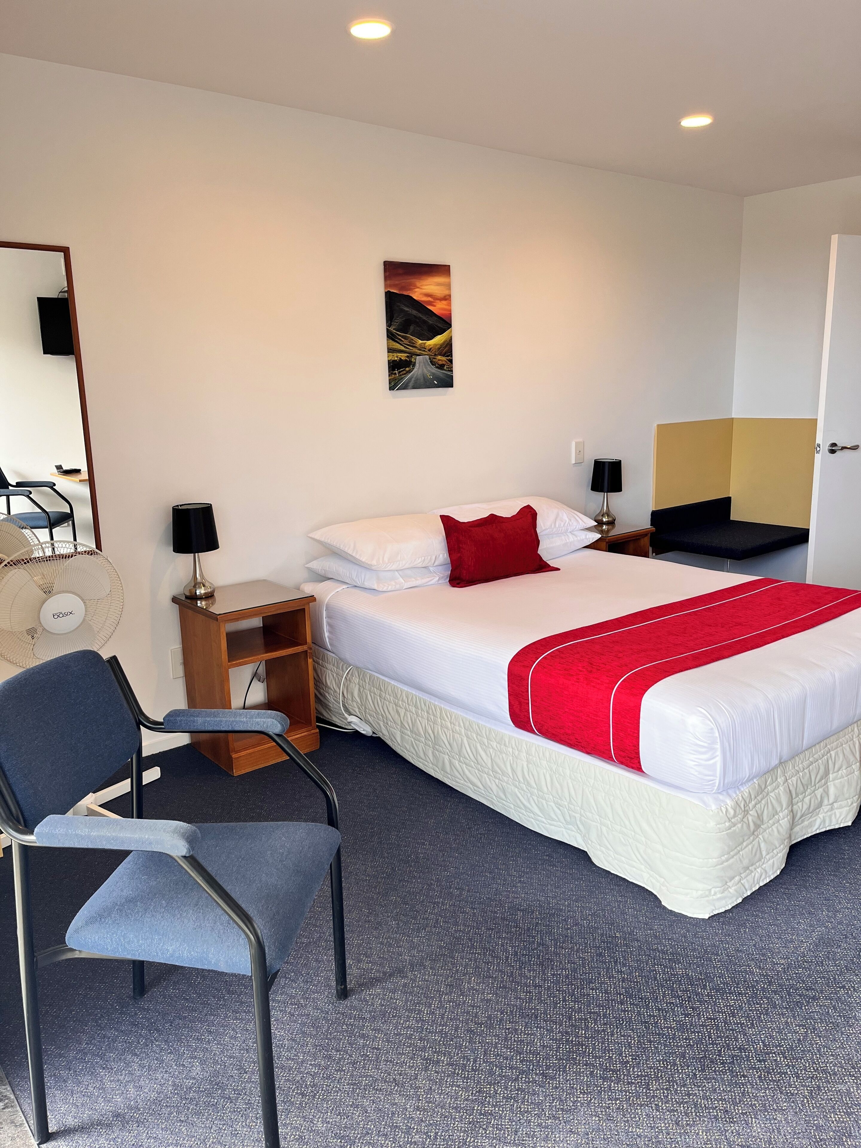 Una habitación de hotel con cama, escritorio, silla, ventilador de pie, un cuadro en la pared y una puerta.