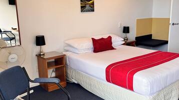 Una habitación de hotel con cama, escritorio, silla, ventilador de pie, un cuadro en la pared y una puerta.