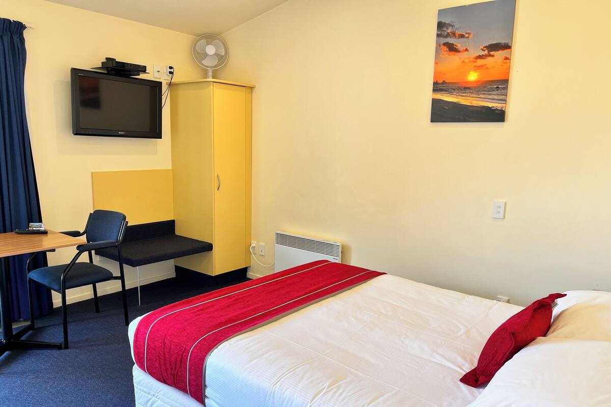 A hotel room with a bed, a desk, a chair, a TV, and a painting of a sunset.