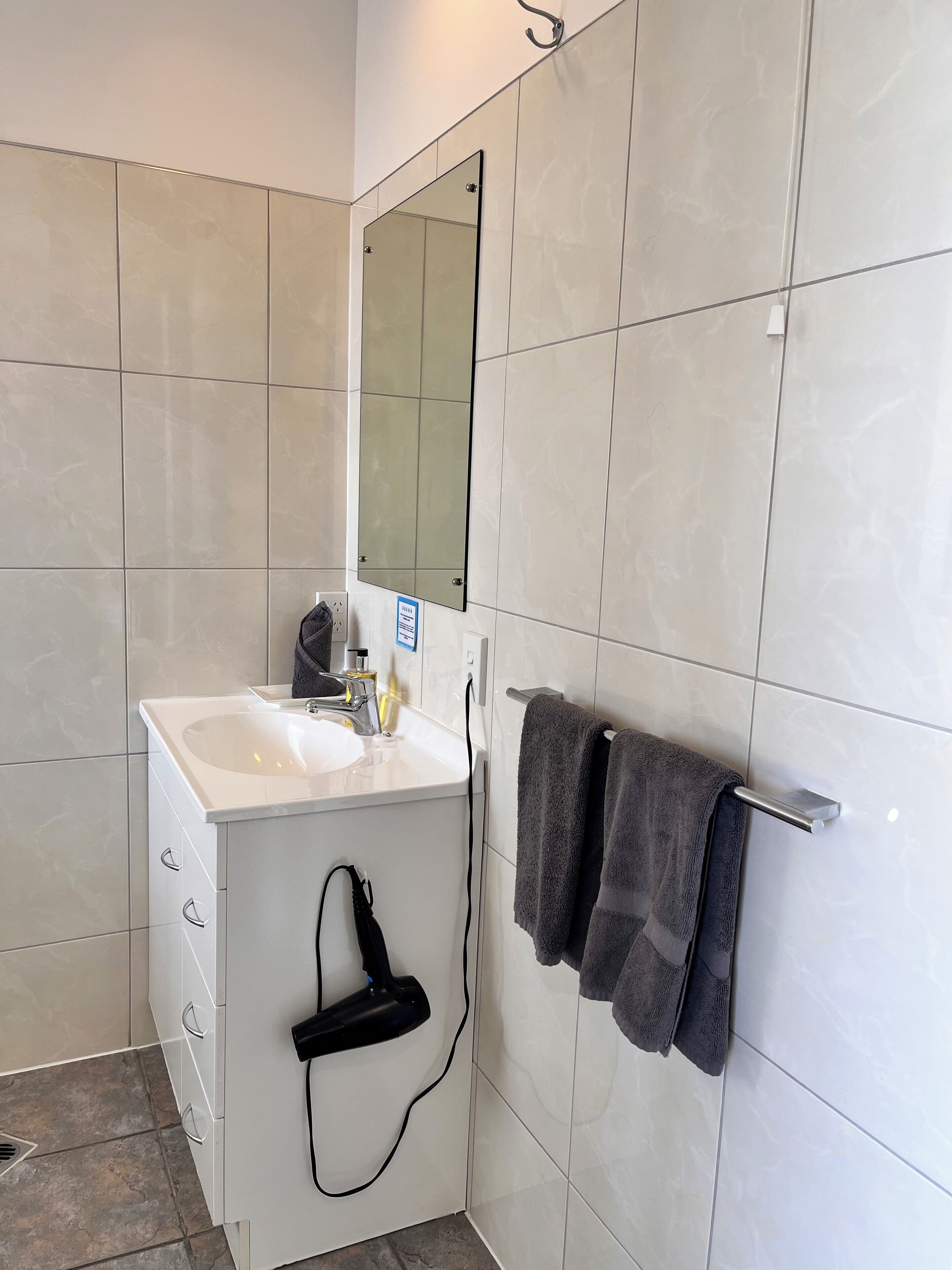 A bathroom with white tiles, a white sink, a mirror, a towel rack with towels, and a black hairdryer.
