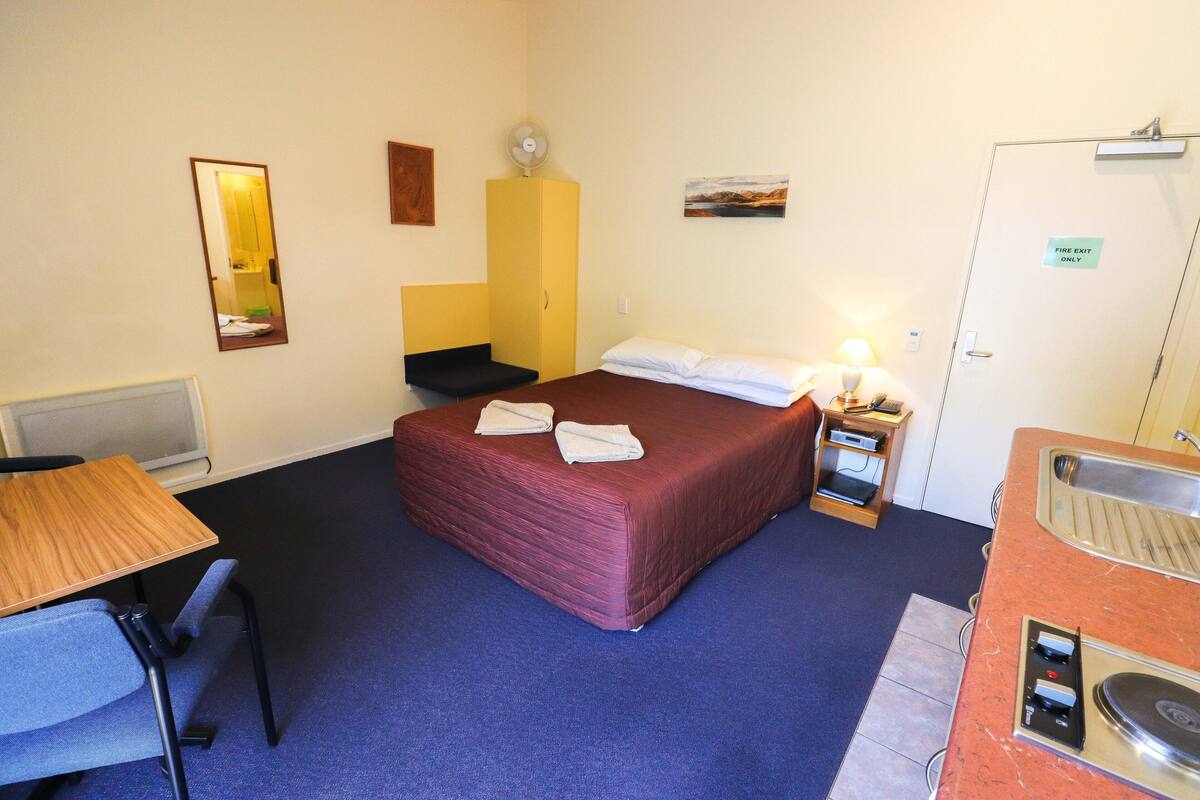 A hotel room with a bed, a desk, a chair, a small table, a sink, and a mirror.
