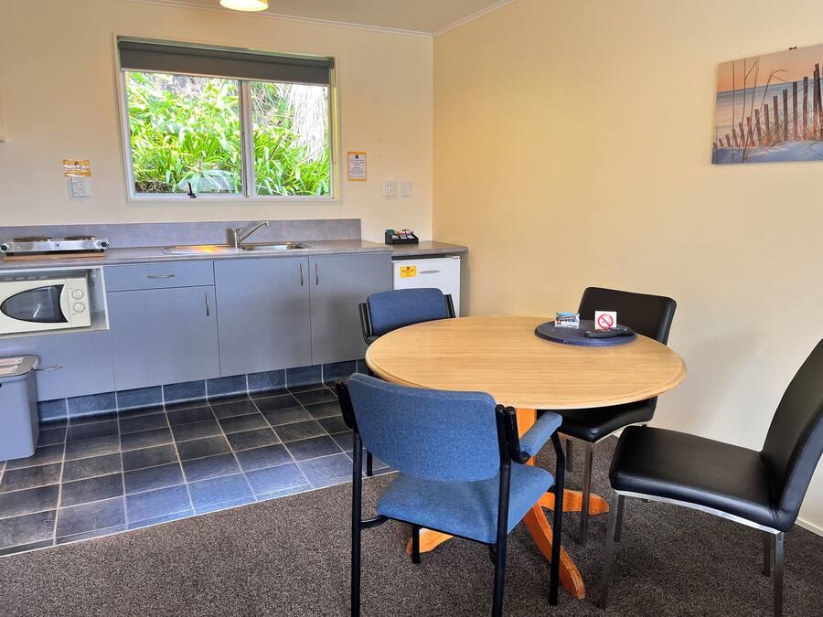 A compact kitchen with a sink, microwave, and stove, a round wooden table with chairs, and a window with a view of greenery.