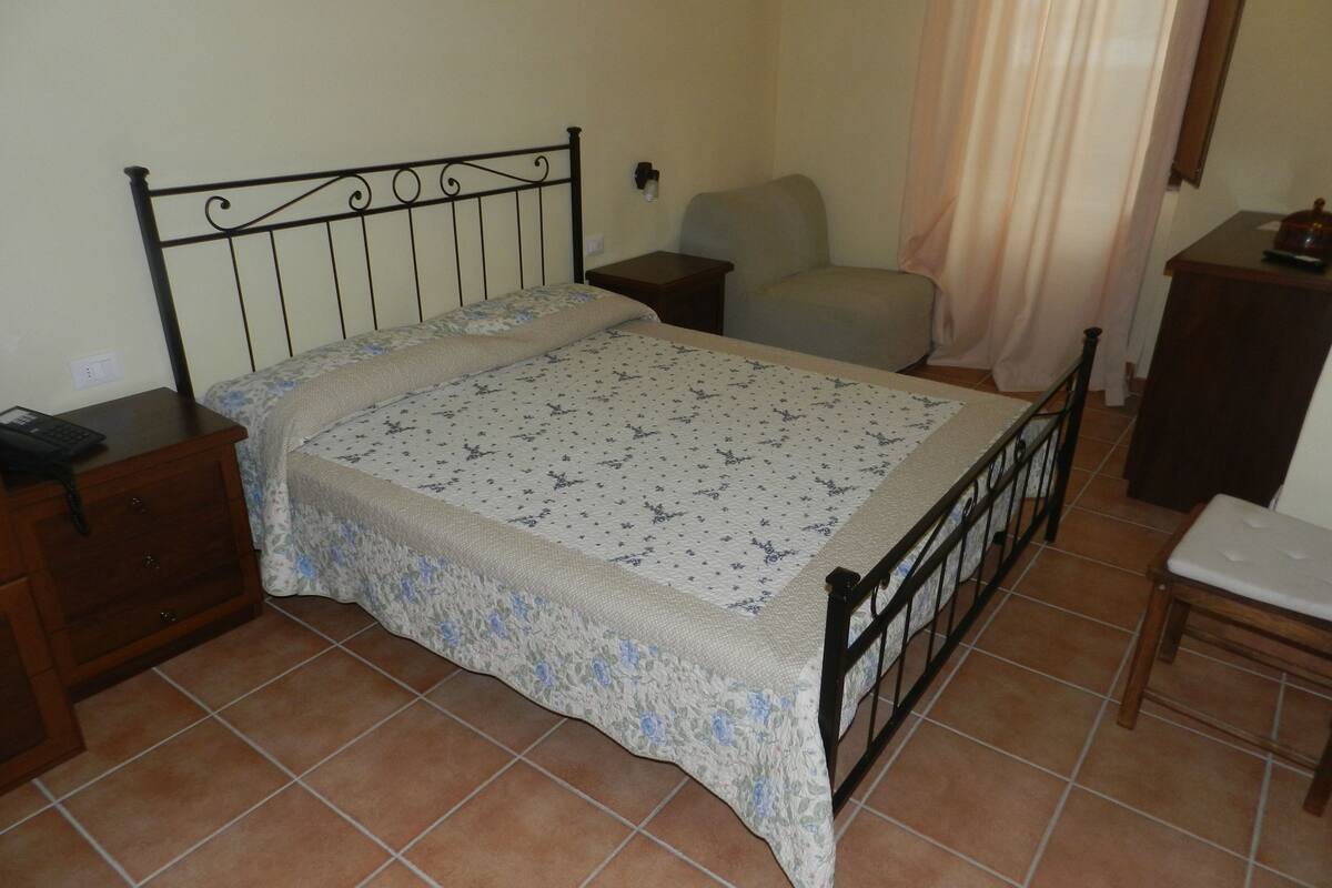 A bedroom with a bed, a nightstand, a chair, a small table, and a curtain.