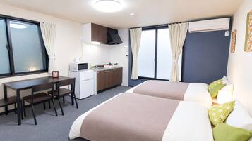 A hotel room with two beds, a kitchenette, a desk, and a window with curtains.