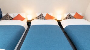 Habitación de hotel con tres camas, cada una con cubrecama azul y almohadas blancas. Hay dos mesitas de noche con lámparas entre las camas.