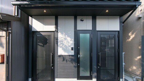 Una entrada moderna de edificio con dos puertas de metal negro, una puerta central con un panel de vidrio esmerilado y un pavimento adoquinado.