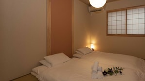 Ein ordentlich gemachtes Bett mit weißen Leinen und aufgerollten Handtüchern, eine Nachttischlampe und ein Fenster mit Gittermuster.
