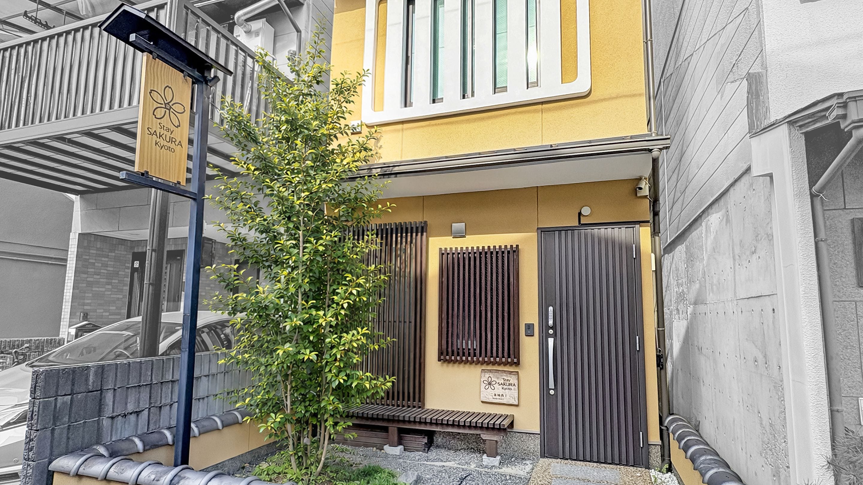 Un bâtiment à la façade jaune, un banc en bois, un petit arbre et un portail en métal.