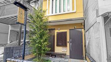 Un bâtiment à la façade jaune, un banc en bois, un petit arbre et un portail en métal.