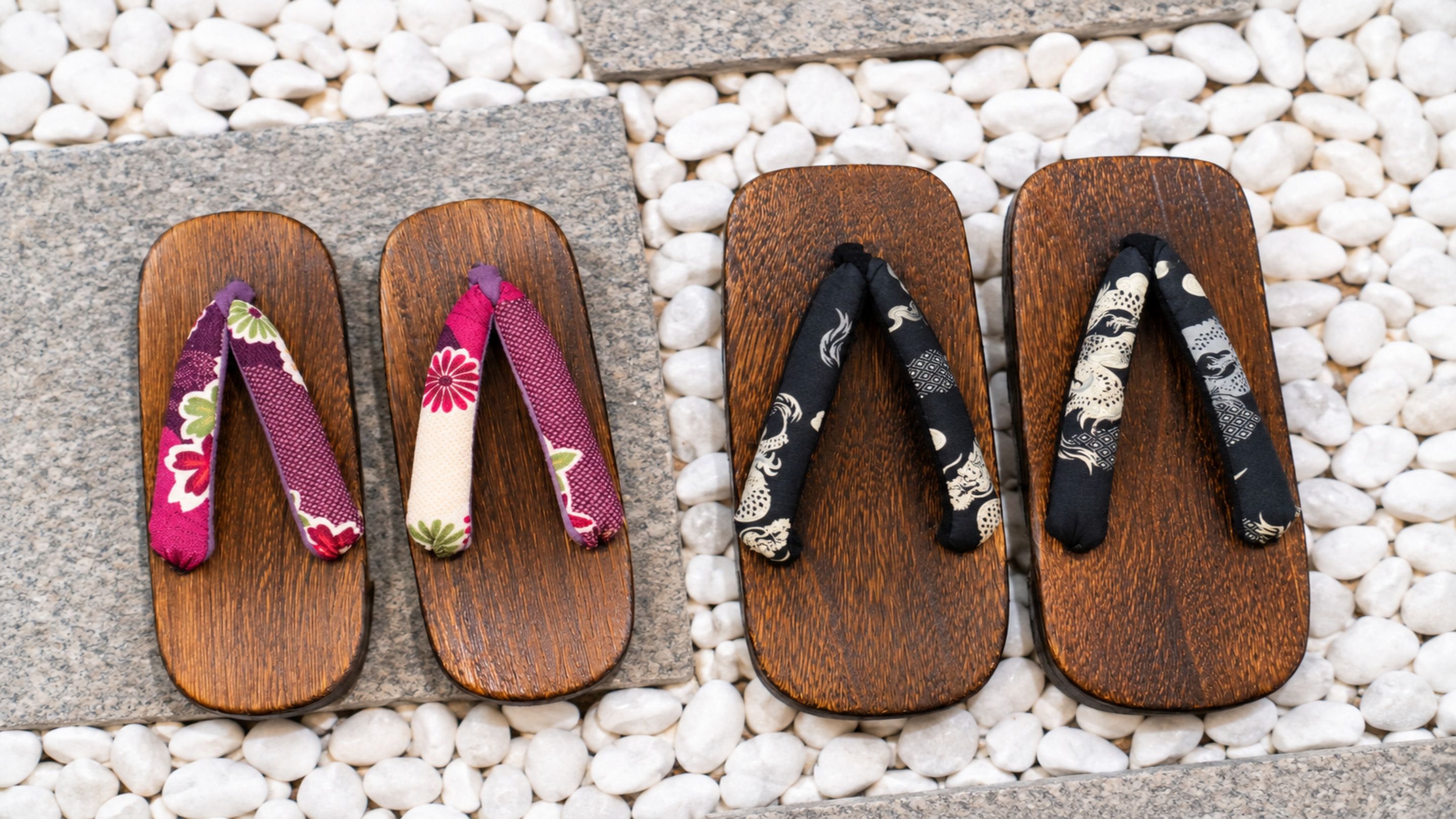 Three pairs of wooden flip-flops with different colored and patterned straps placed on a bed of white pebbles.