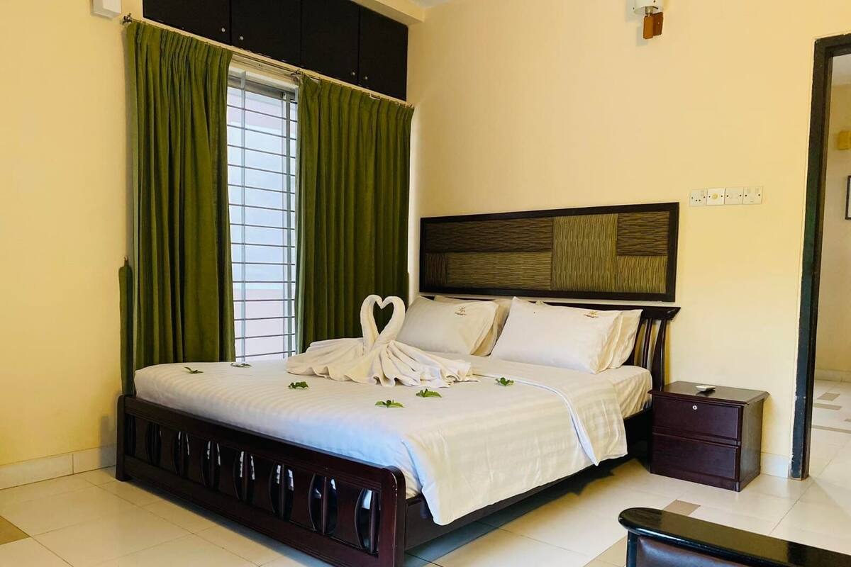 A neatly made bed with a heart-shaped towel arrangement, a wooden headboard, and a nightstand with a lamp.