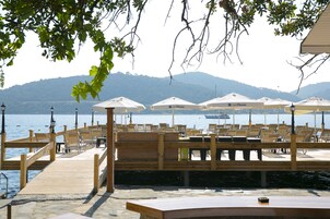 A wooden pier with lounge chairs and umbrellas, overlooking a calm body of water with boats and mountains in the background.