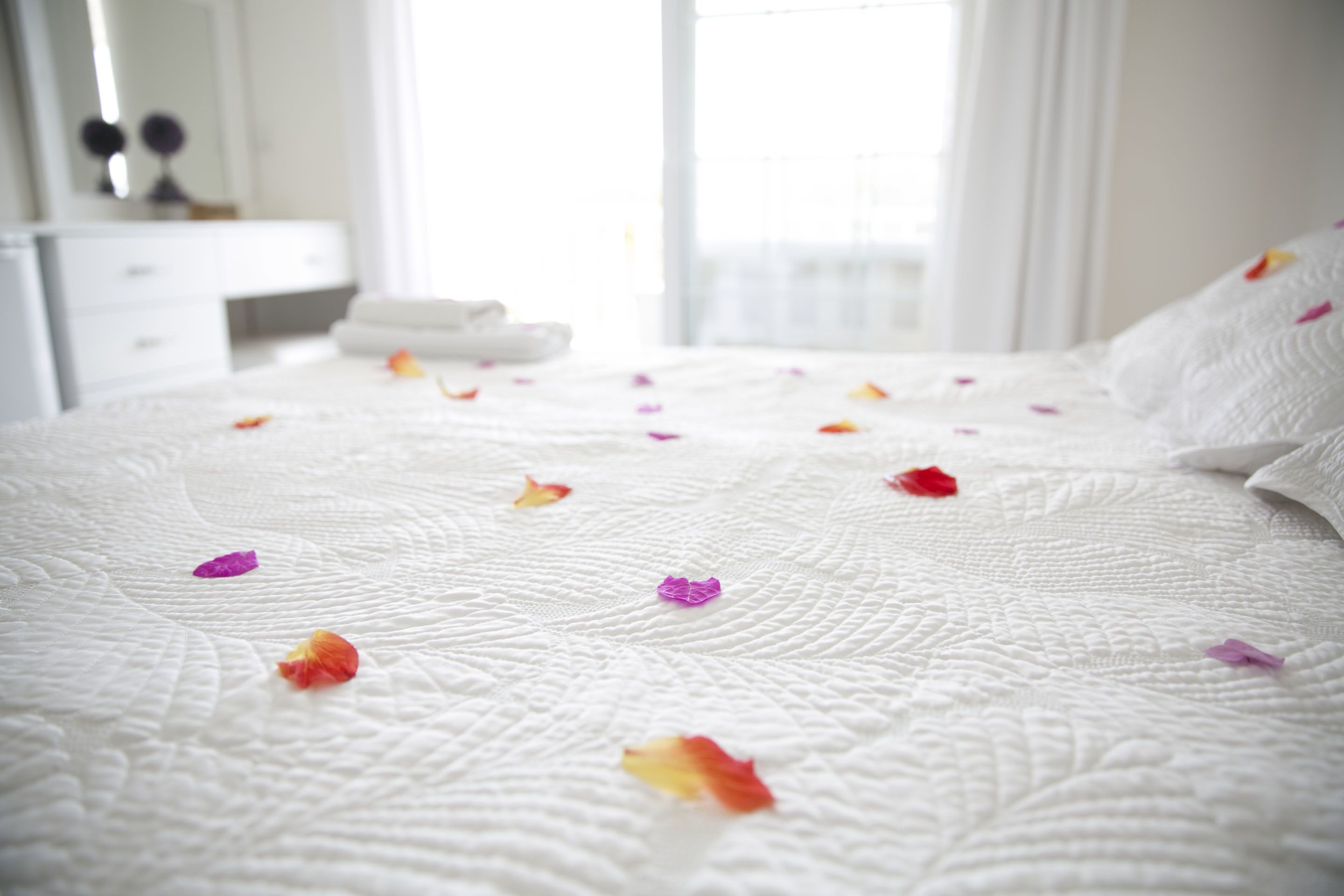 Una cama bien hecha con colcha blanca y pétalos de flores esparcidos.