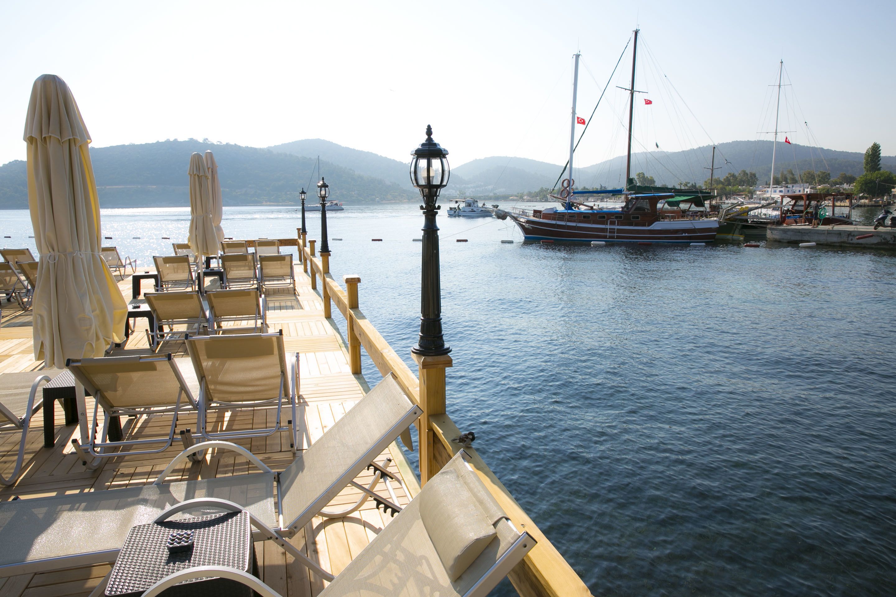Terraza con tumbonas y una mesa, con vistas a un agua tranquila con barcos y una cadena montañosa a lo lejos.