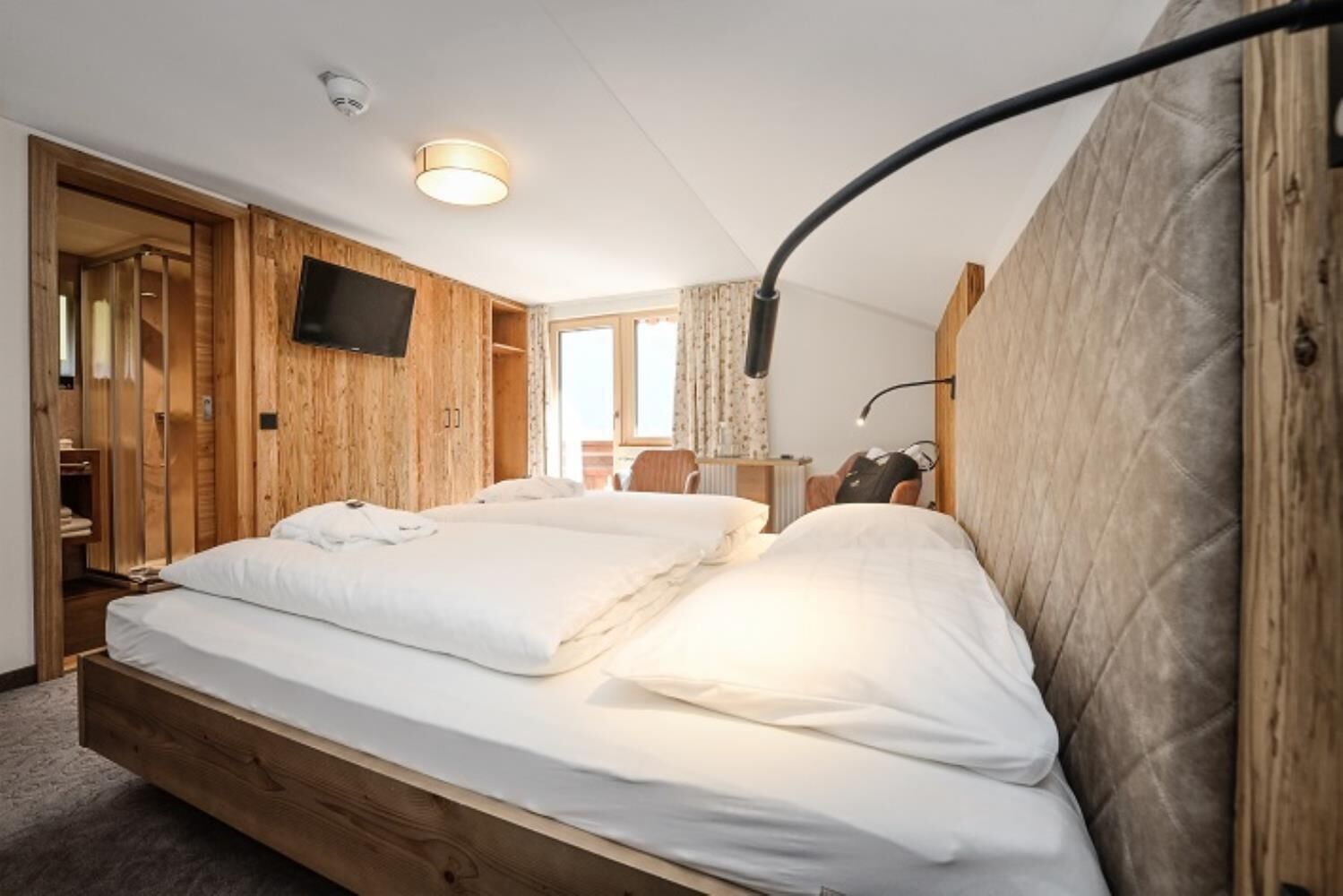 Ein Doppelbett mit weißer Bettwäsche, ein hölzerner Kopfteil, ein Flachbildfernseher an der Wand und eine Deckenleuchte.