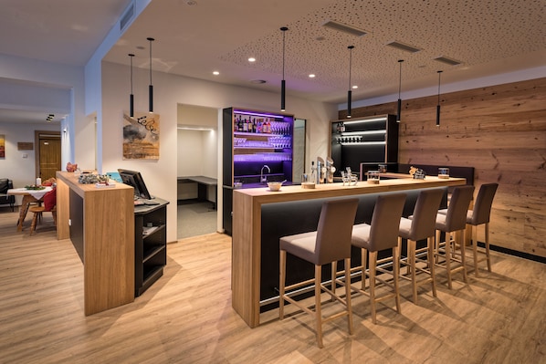 A modern bar area with a wooden bar counter, bar stools, and a wine cooler. - Hotel Alpenfeuer Montafon (Sankt Gallenkirch)