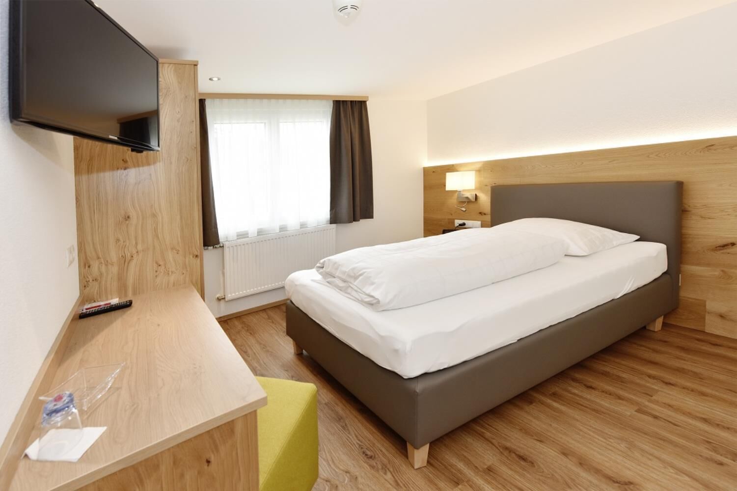A hotel room with a bed, a desk, a TV, a window with curtains, and a bedside lamp.