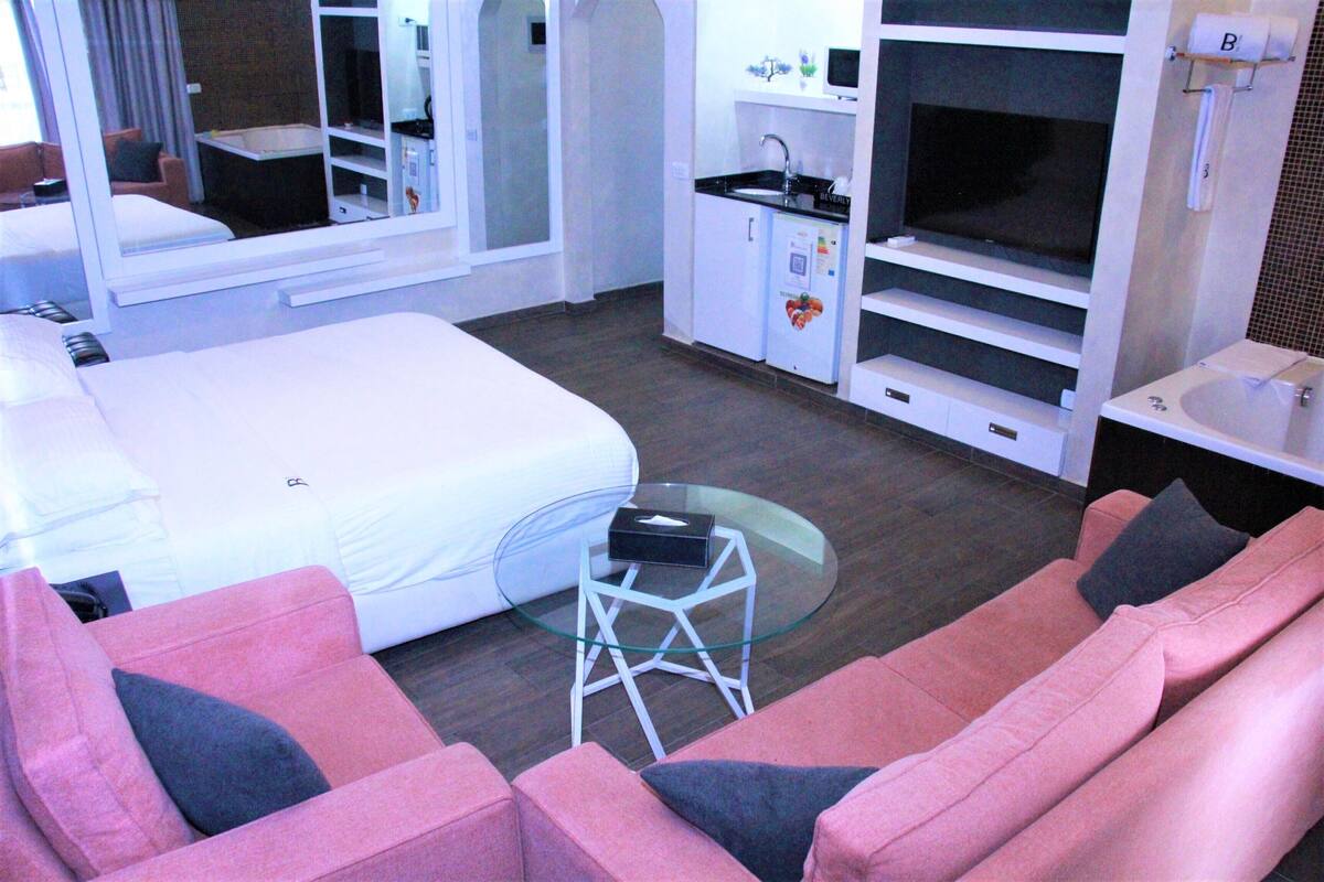 A compact hotel room with a bed, a sofa, a small table, a TV, a mini-fridge, and a sink.