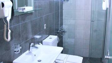 Casa de banho com sanita, lavatório e chuveiro. Secador de cabelo na parede e um porta-toalhas com uma toalha.