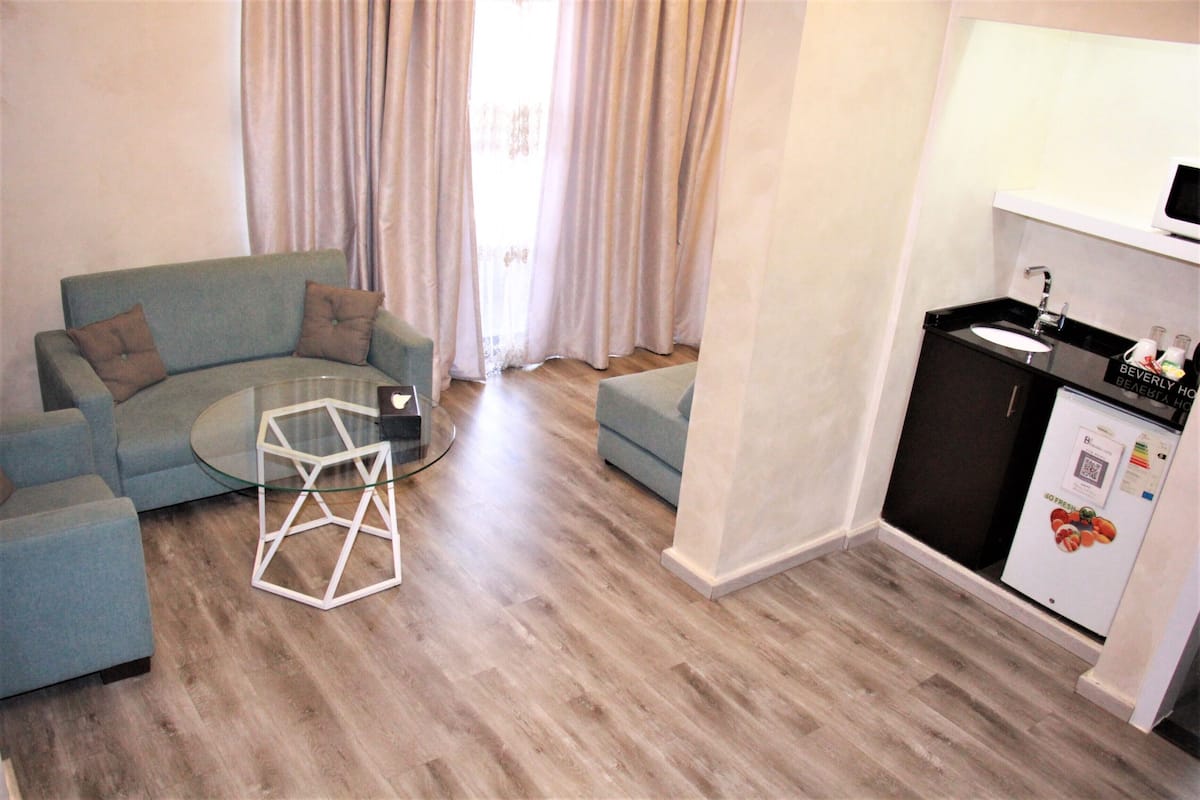 A living room with a blue sofa, a glass coffee table, and a kitchen area with a refrigerator and sink.