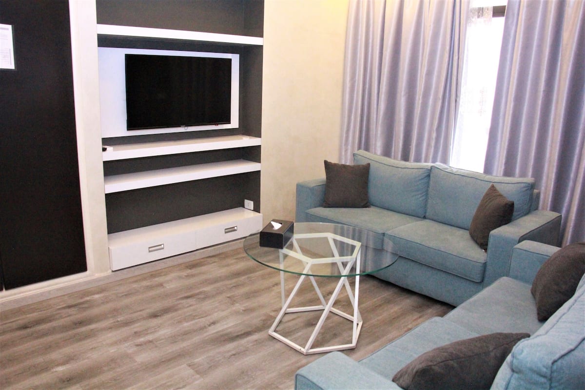 A living room with a TV on a white shelf, a glass coffee table, and a light blue sofa with cushions.
