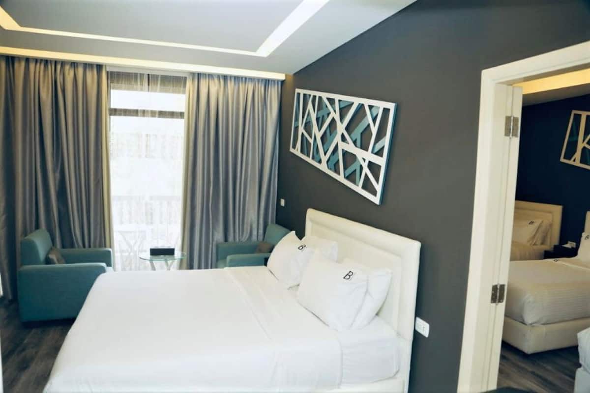 A hotel room with a large bed, two armchairs, a decorative wall piece, and a view of the city through the window.