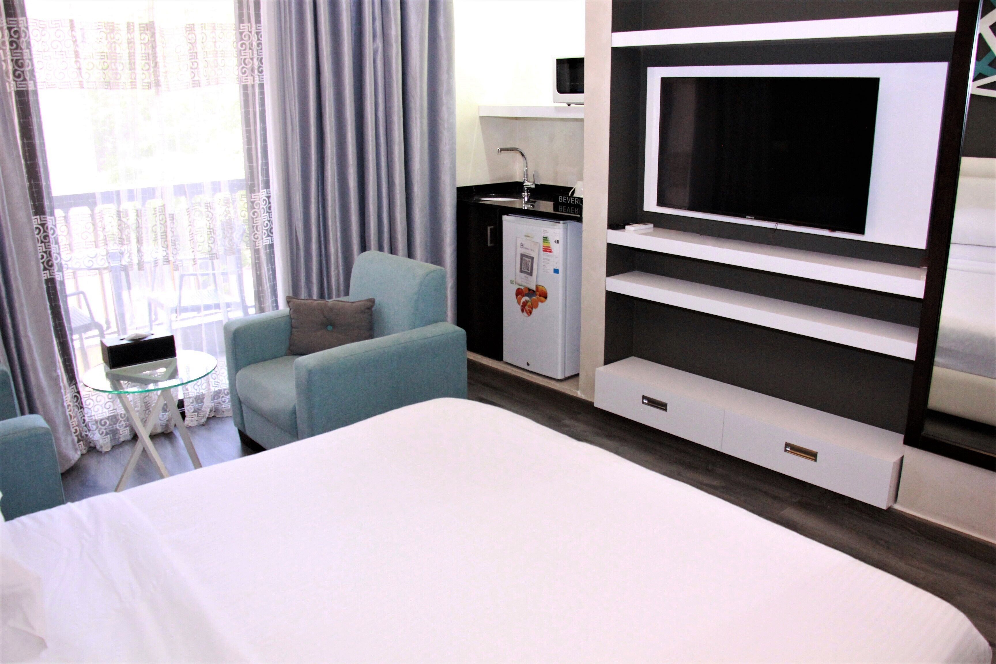Habitación de hotel con cama, zona de estar con sillas, una mesita, refrigerador, televisor y ventana con cortinas.