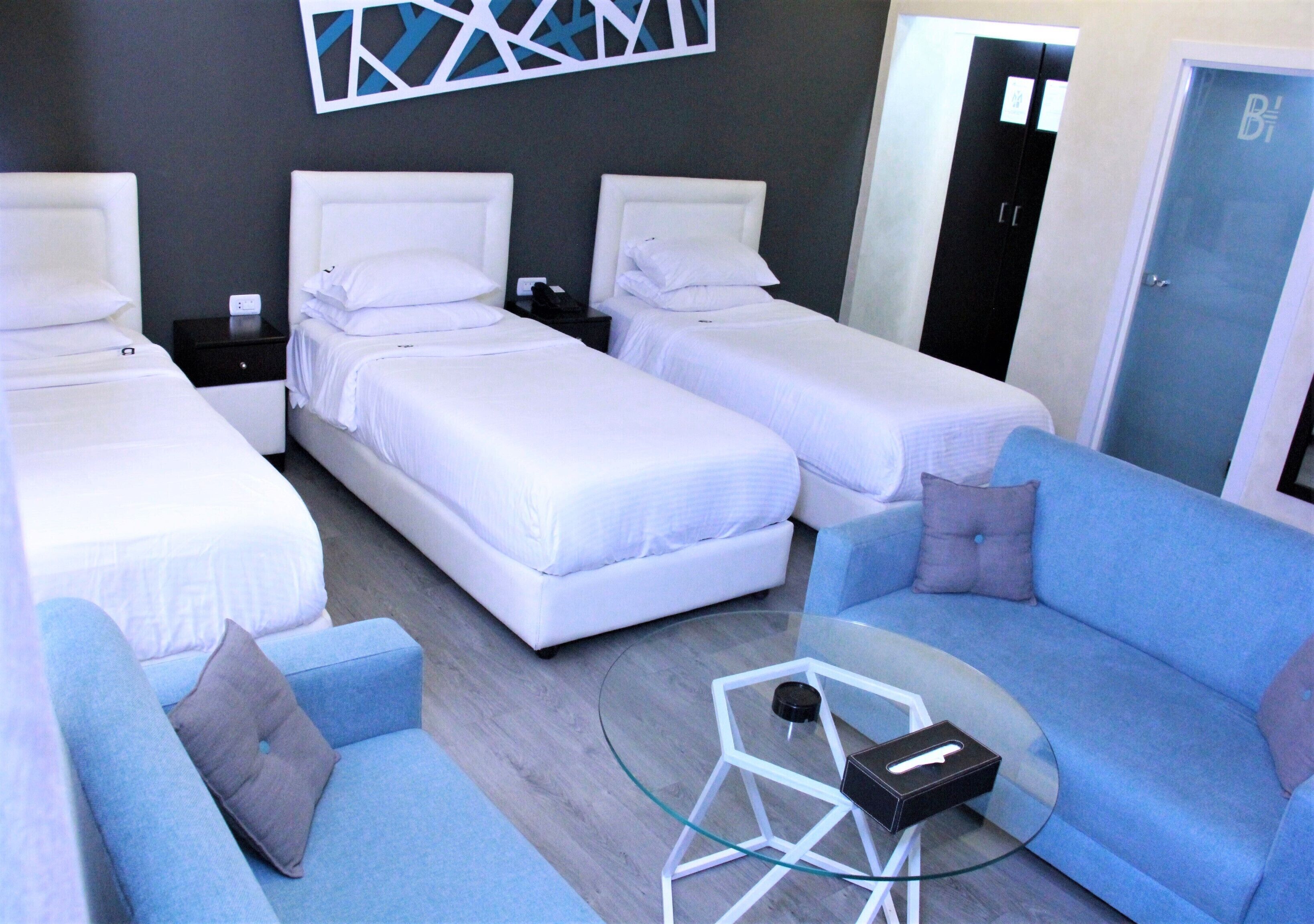 Habitación de hotel con tres camas, un sofá azul, una mesa de centro de vidrio y un cuadro abstracto moderno en la pared.