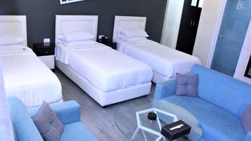 Habitación de hotel con tres camas, un sofá azul, una mesa de centro de vidrio y un cuadro abstracto moderno en la pared.