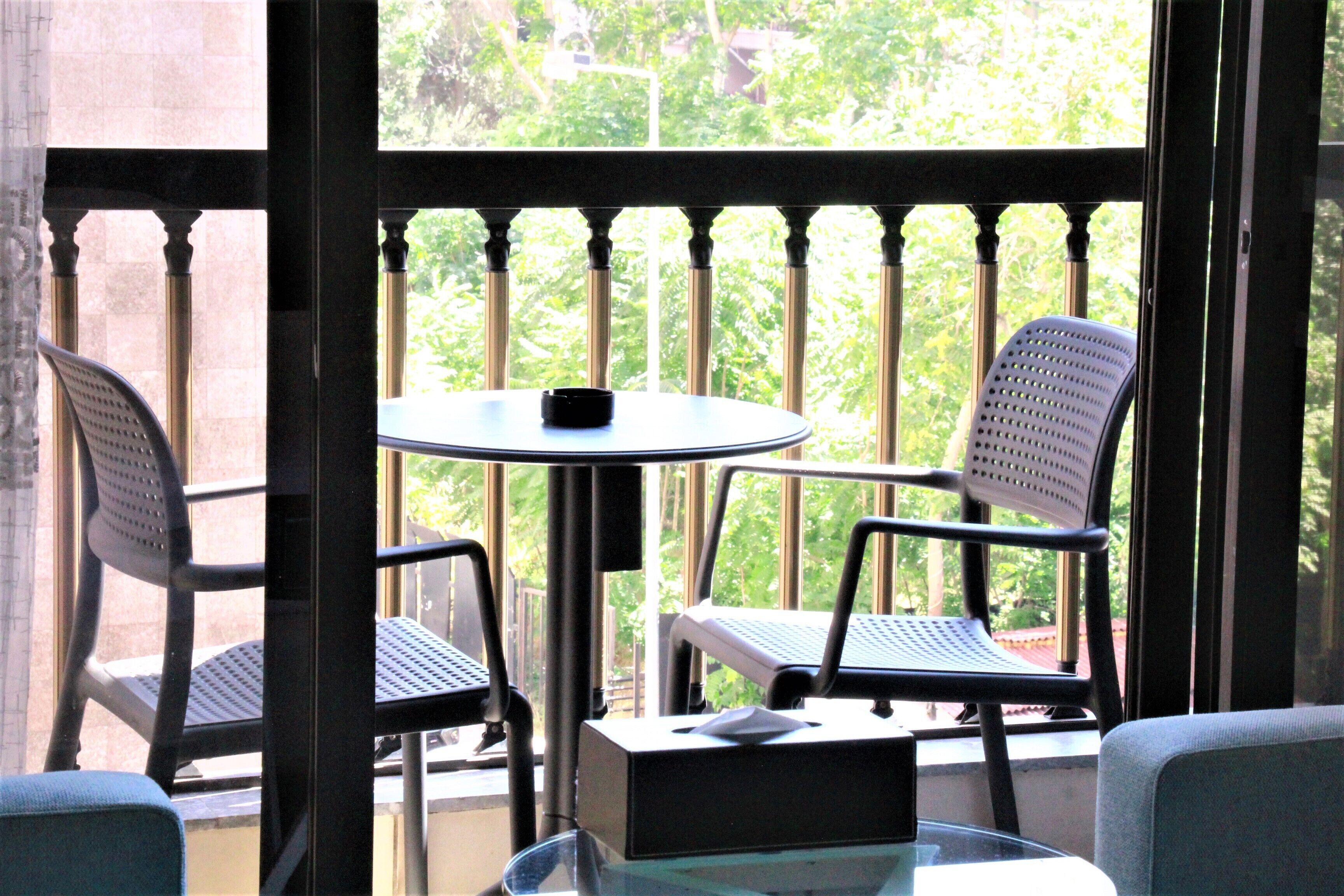 A balcony with a round table and two chairs, a small table with a black object, and a view of greenery through the railing.